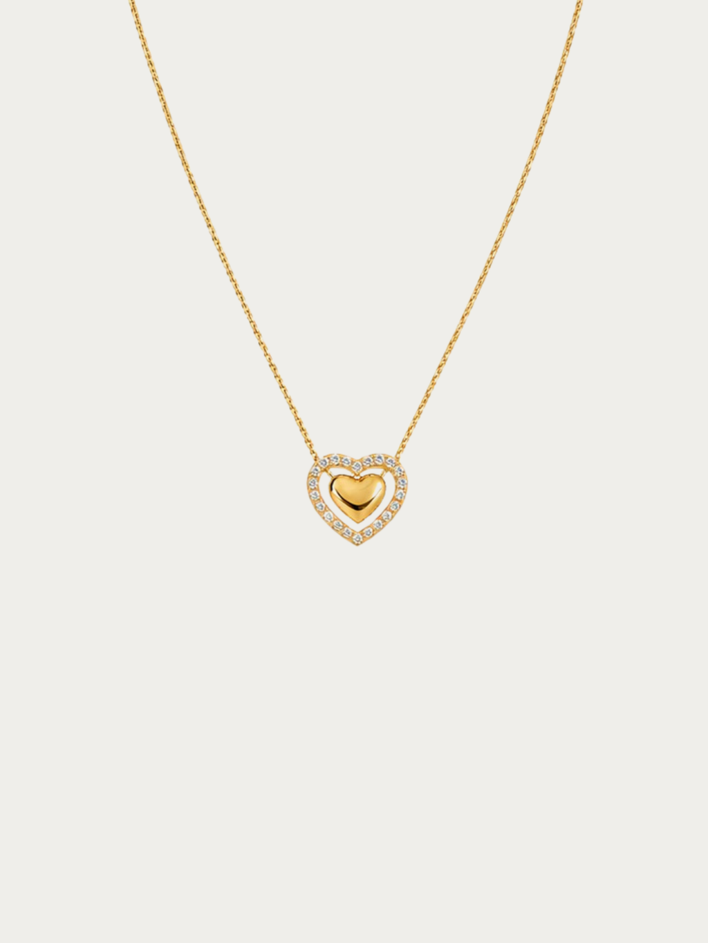 A delicate gold chain necklace featuring a heart-shaped pendant with a polished gold center heart surrounded by a halo of small clear stones, set against a solid light gray background.