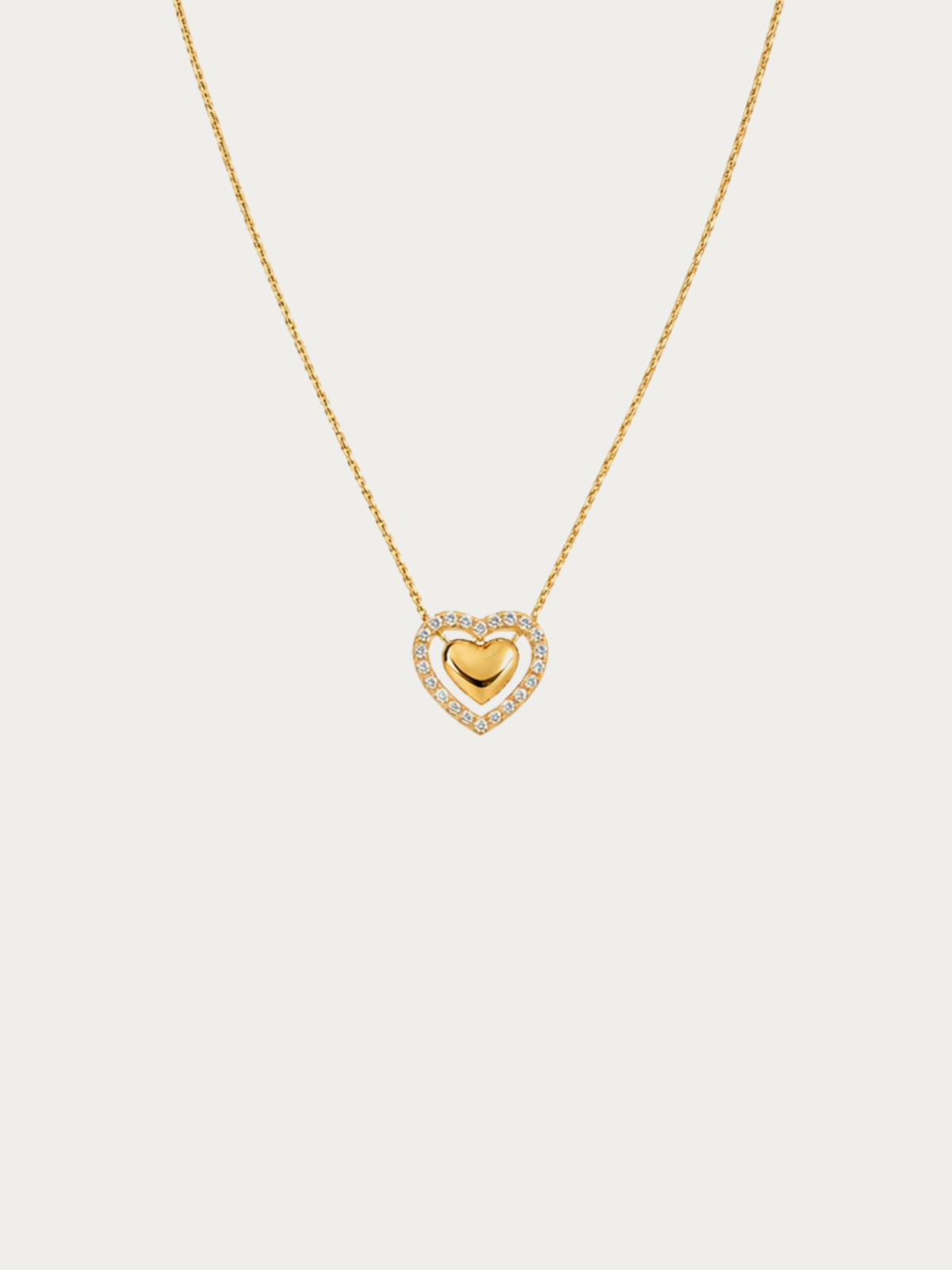 A delicate gold chain necklace featuring a heart-shaped pendant with a polished gold center heart surrounded by a halo of small clear stones, set against a solid light gray background.