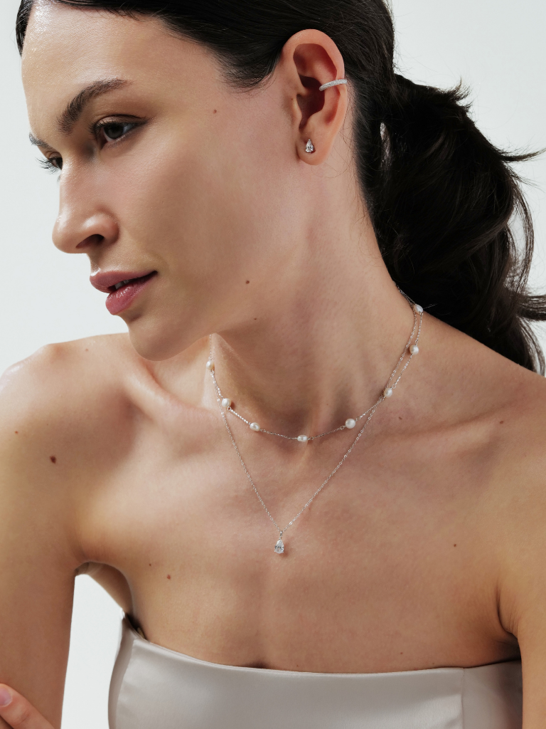 A close-up of a woman wearing a delicate double-strand pearl necklace with a small silver pendant and matching stud earrings against a light gray background.